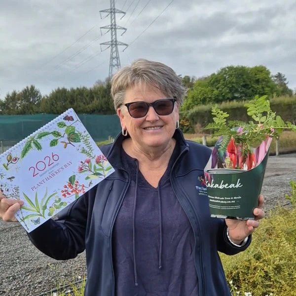 Marie from Plant Hawke's Bay holding the Plant Hawke's Bay Botanical Calendar 2026 and a young Kakabeak plant